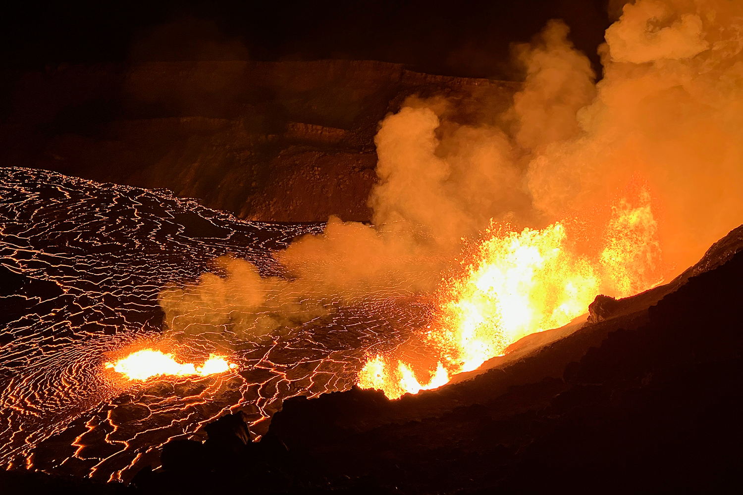 Nighttime view in which lava and smoke escape from the ground and lava is shown in cracks in the ground.