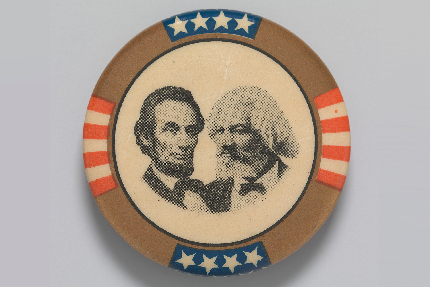 A round pin with stars and stripes around the edges has a composite of portraits of Abraham Lincoln and Frederick Douglass in the center.