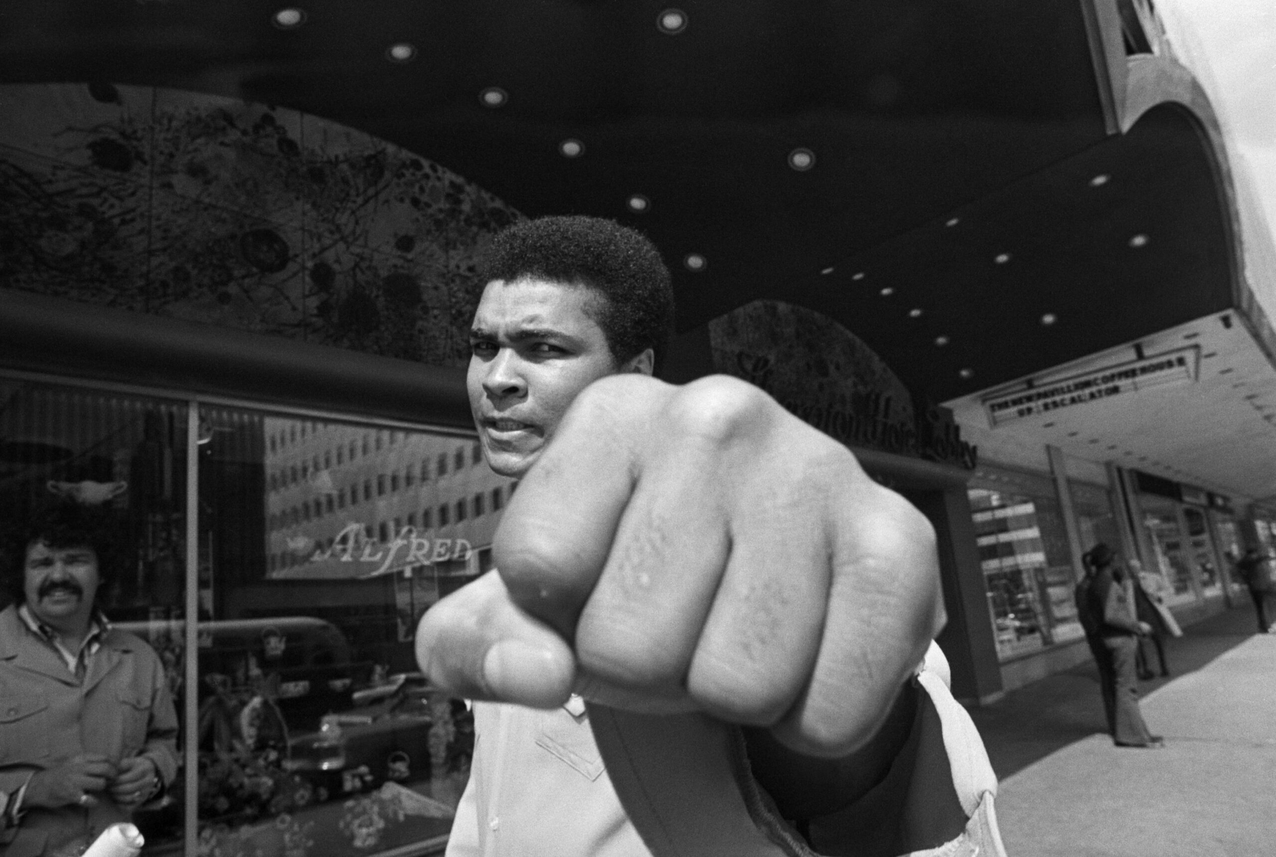 Muhammad Ali stands on a sidewalk and holds his fist to the camera as if punching it.