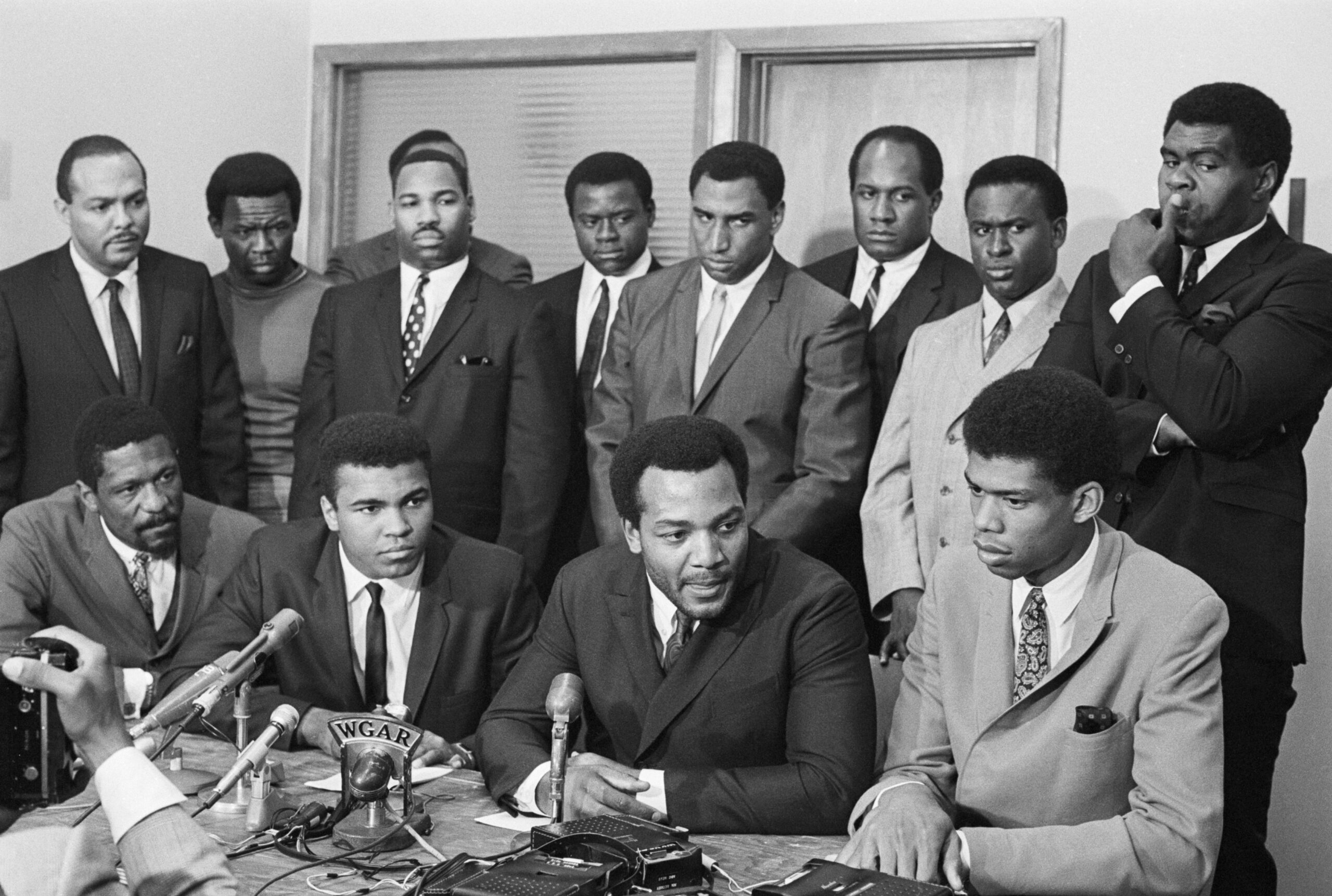 Muhammad Ali sits at a table where microphones have been set up and is surrounded by other athletes.