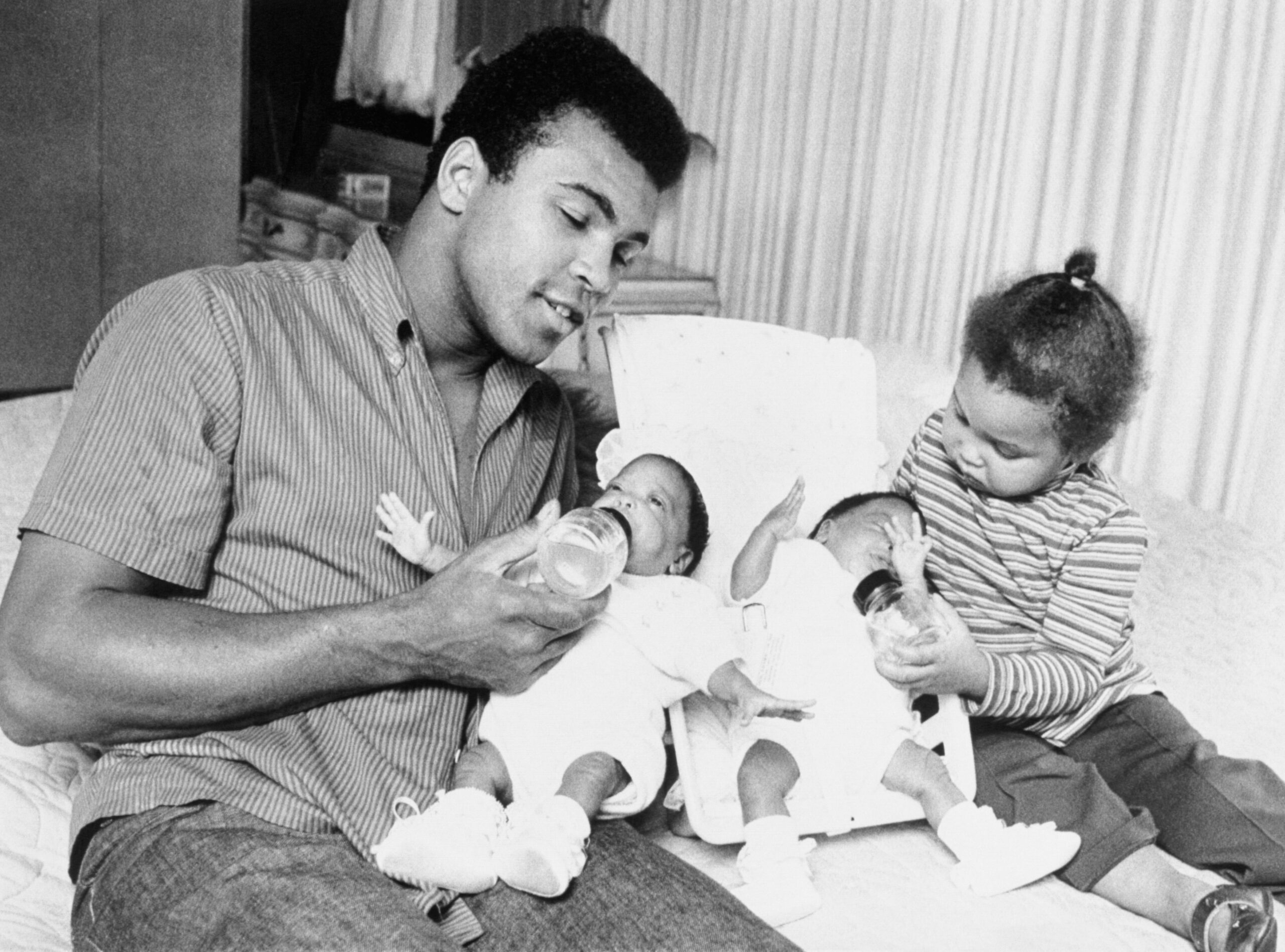 Young Muhammad Ali sits on a couch and feeds one of his infant twins while his toddler plays with the other twin.