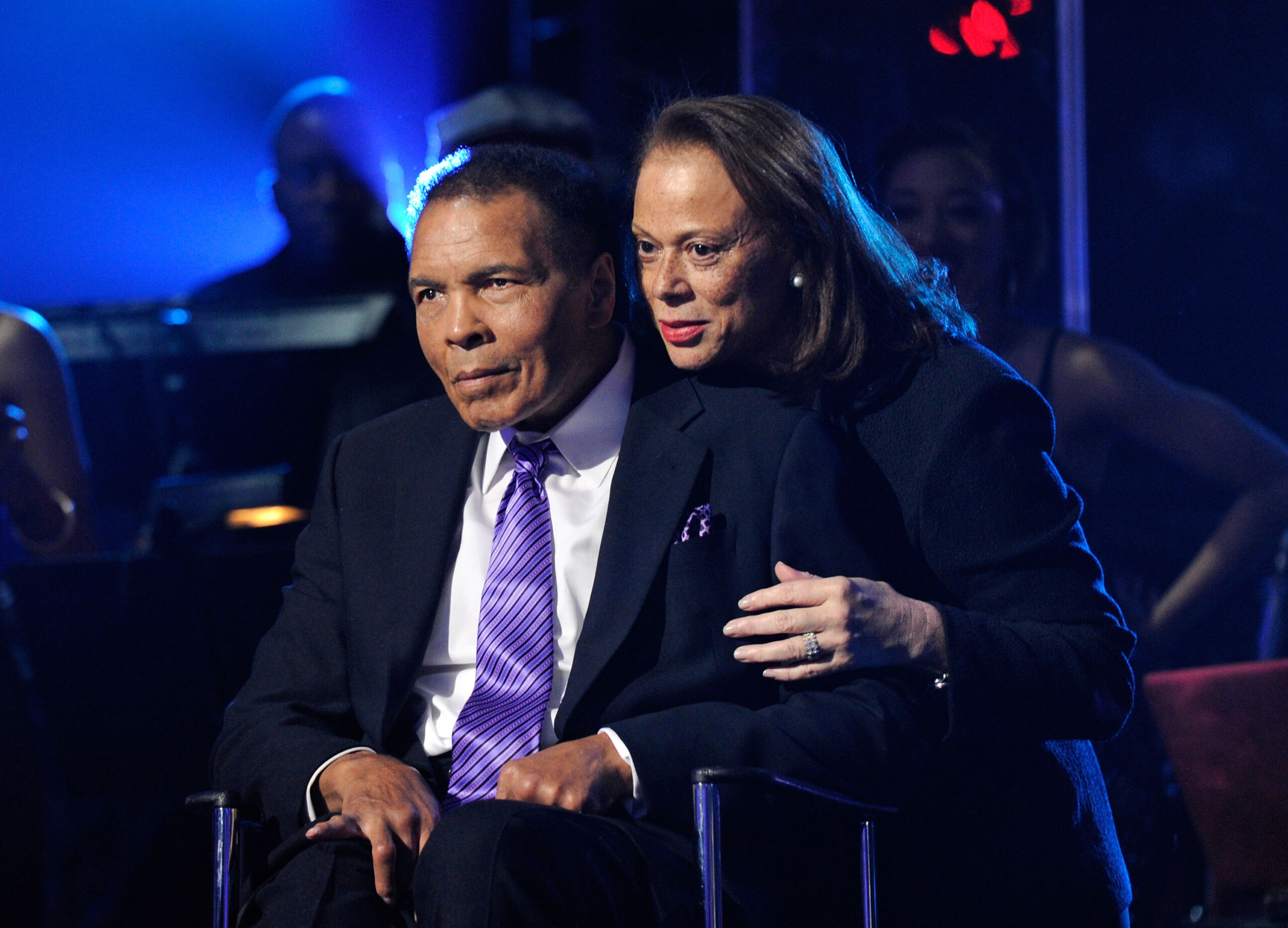 Older Muhammad Ali is seated while his wife stands behind him and embraces him.