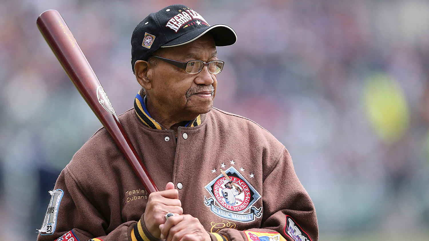 An older Ron Teasley holds a baseball bat against his right shoulder and wears a Negro Leagues jacket and hat.