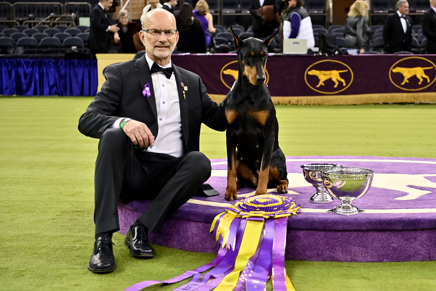 Andy Linton and Penny the dog sit on a raised platform and pose with two silver trophies and a purple and yellow ribbon.