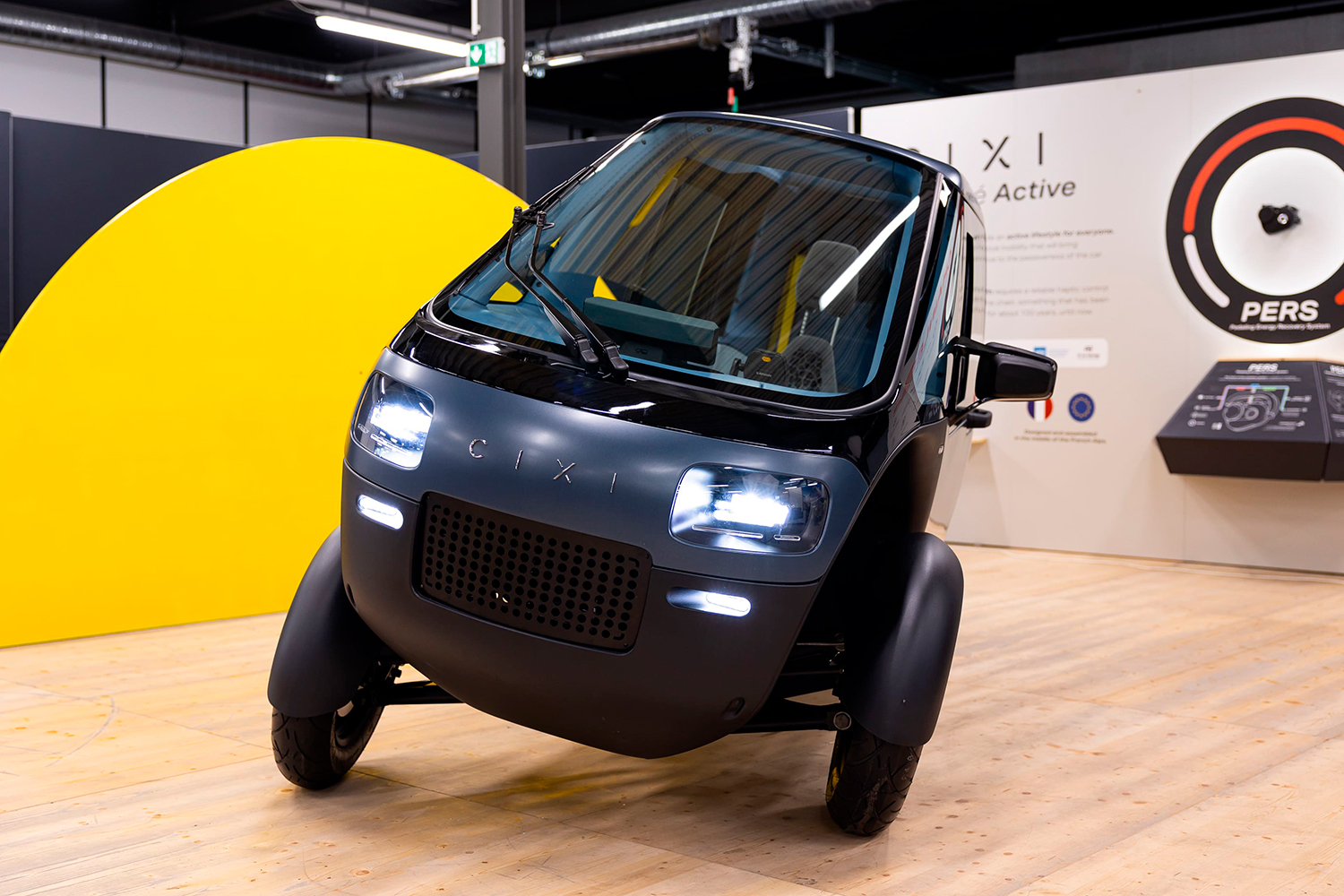 A small black three wheeled vehicle with its headlights on sits in a showroom in front of a display.