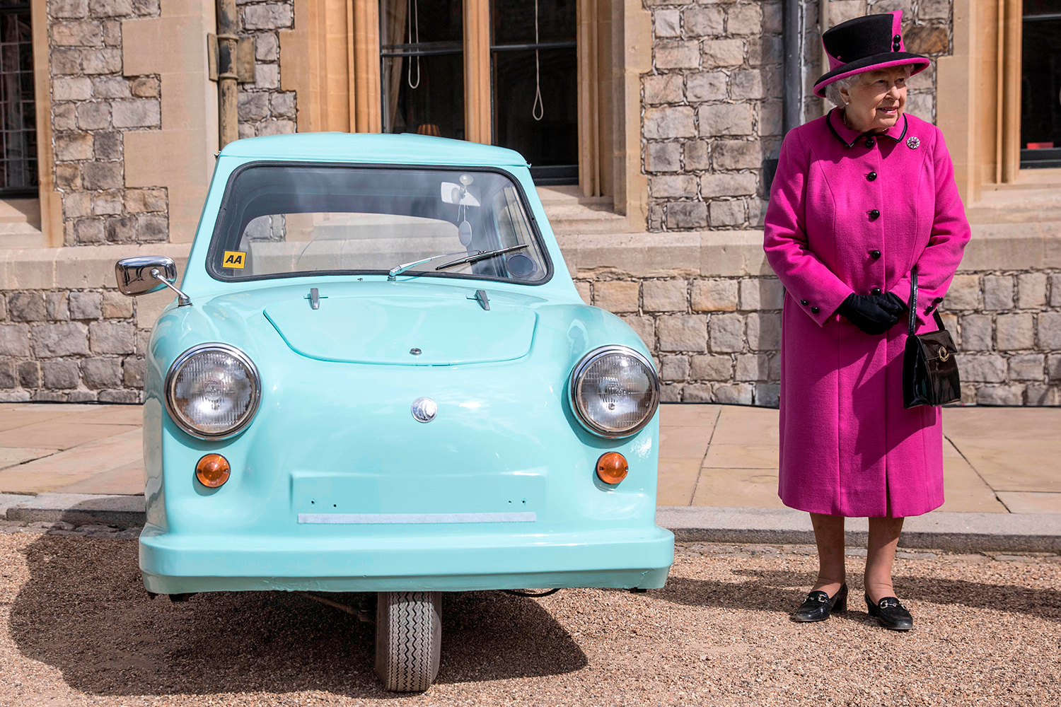 Queen Elizabeth II stands next to a blue Invacar that is parked on the street.