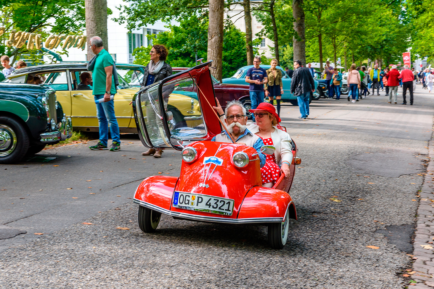 A man and a woman ride a tiny car through an area where other classic cars are parked and people are walking.
