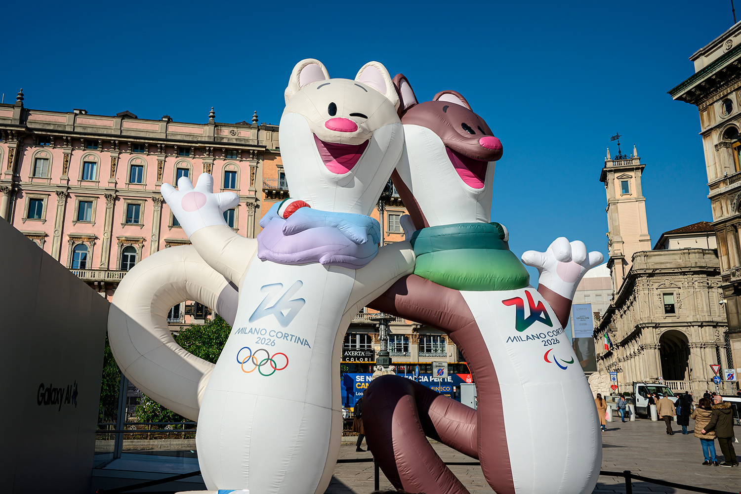Two inflatable stoats with the words Milano Cortina and the Olympic rings on their chests are in an Italian city square.