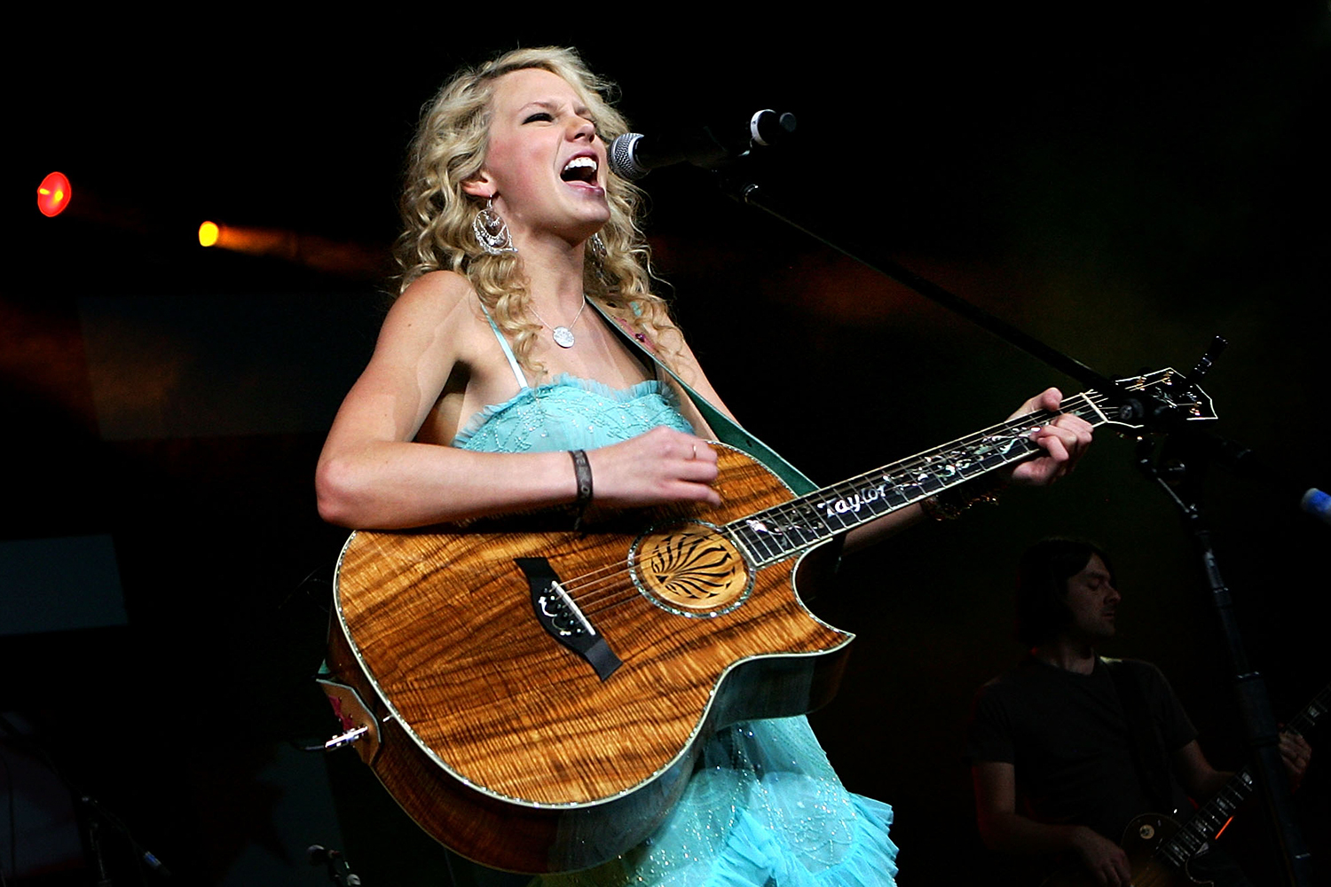 A teenage Taylor Swift plays a guitar and sings into a microphone.