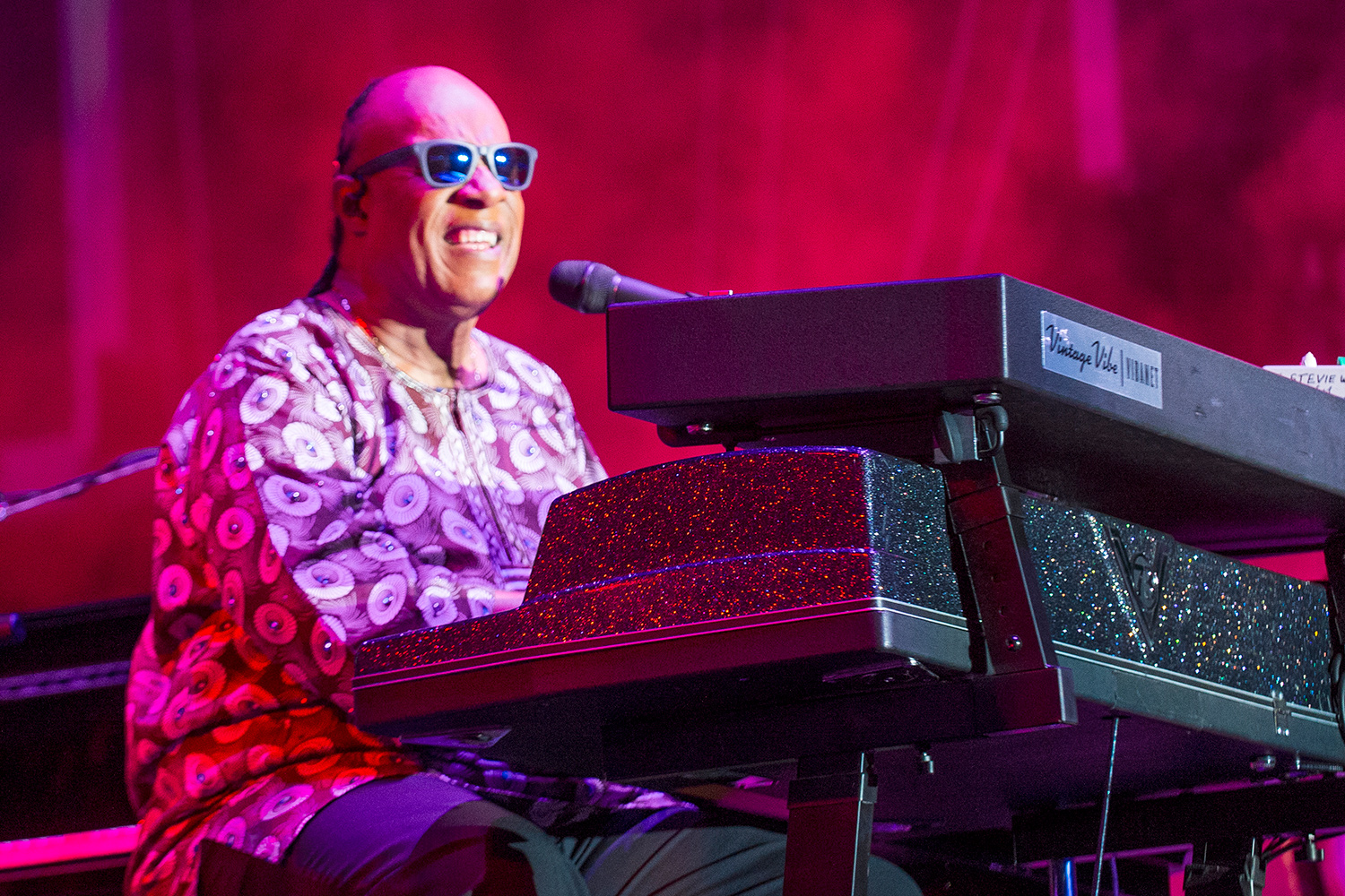 An adult Stevie Wonder sits at a keyboard onstage and smiles.