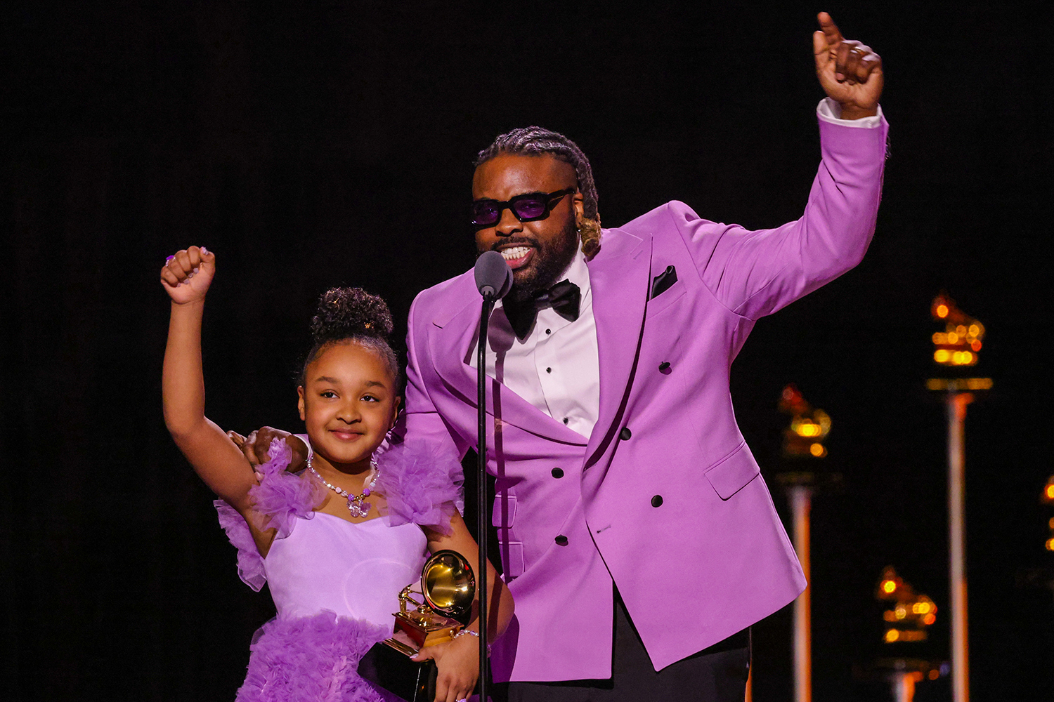 Aura V holds a Grammy Award as she stands at a microphone with her father and they both hold their arms up.