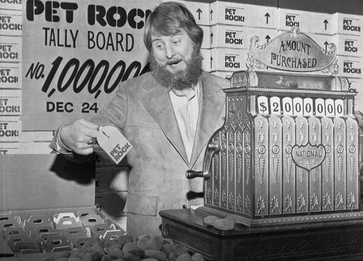 Gary Dahl stands at a cash register and holds a box that reads Pet Rock next to rocks and additional boxes and in front of a sign saying one million rocks have been sold.