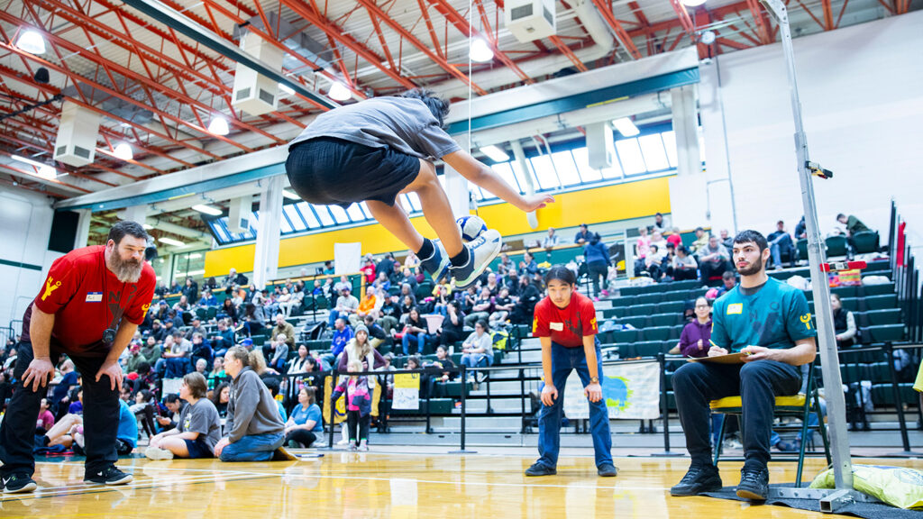 In a gym, a teen has jumped up and is about to kick a hanging ball with two feet as officials and spectators watch.