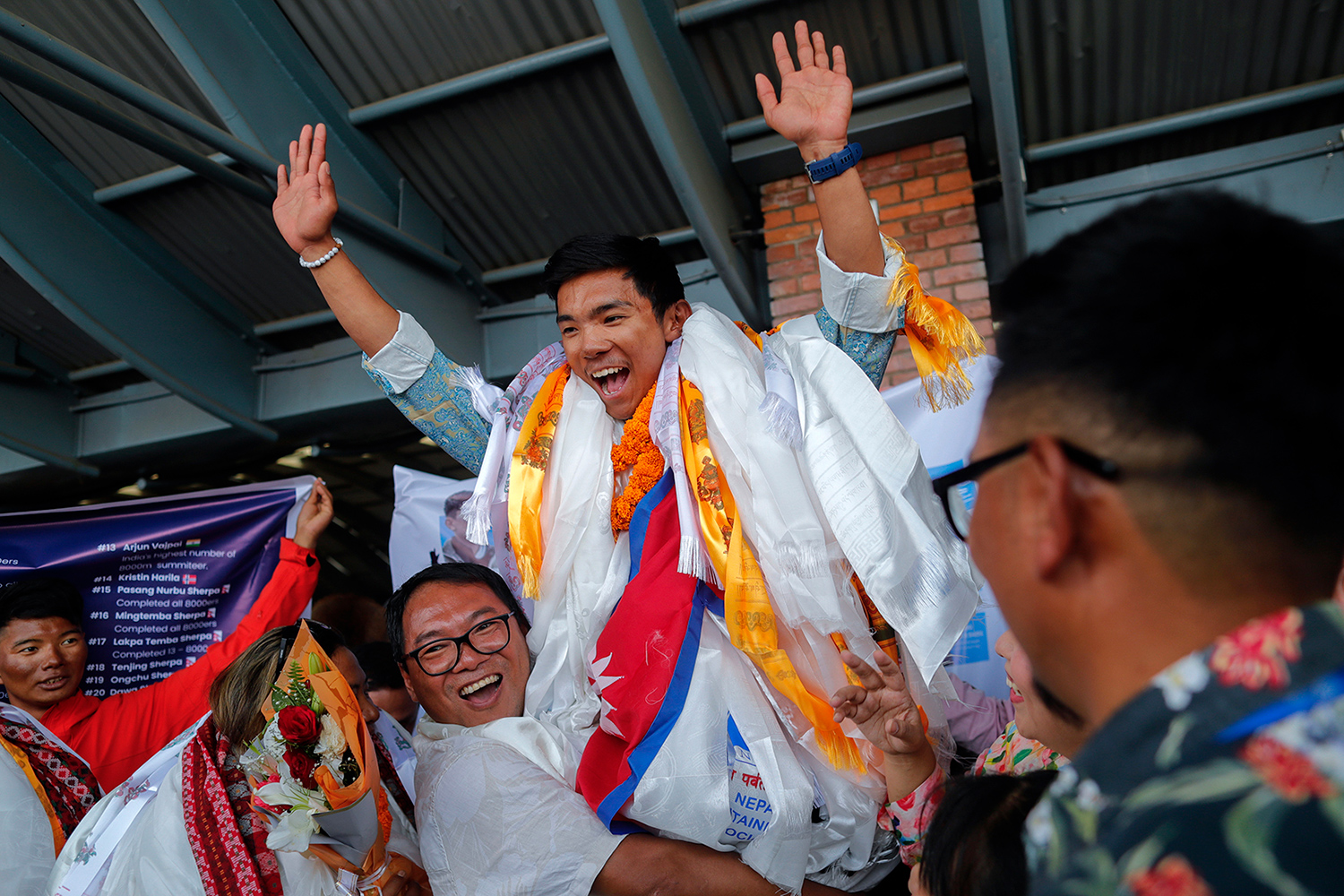 A man holds up Nima Rinji Sherpa, who has both arms up in celebration and is surrounded by a celebrating crowd.