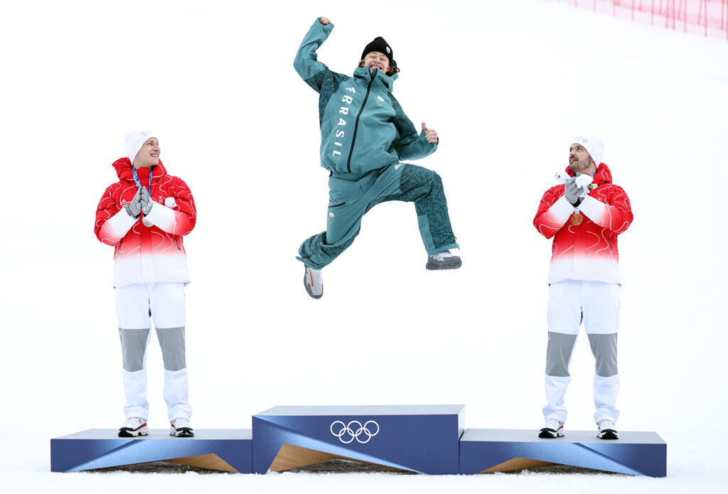 Lucas Pinheiro Braathen is in the air after jumping from the top position on the Olympic podium as silver and bronze winners Marco Odermatt and Loic Meillard watch.