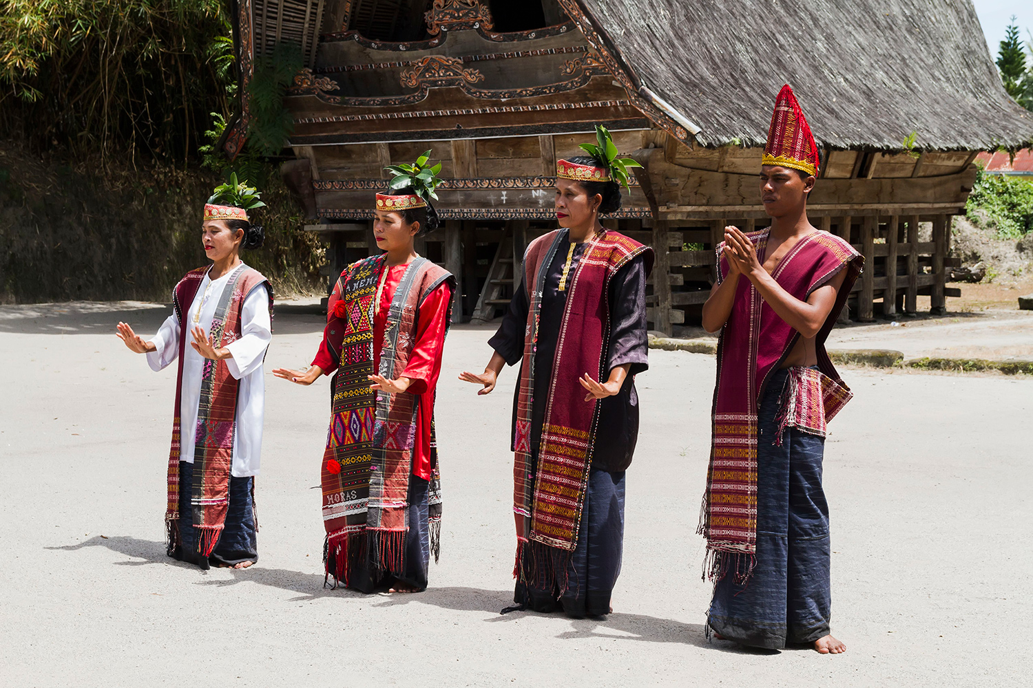 Four people in traditional dress stand in dance poses in front of a traditional Indonesian building.