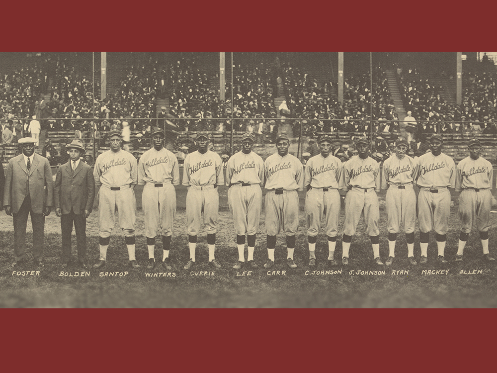 Several players in uniform and two men in suits pose for a photo inside a stadium with spectators in the background.