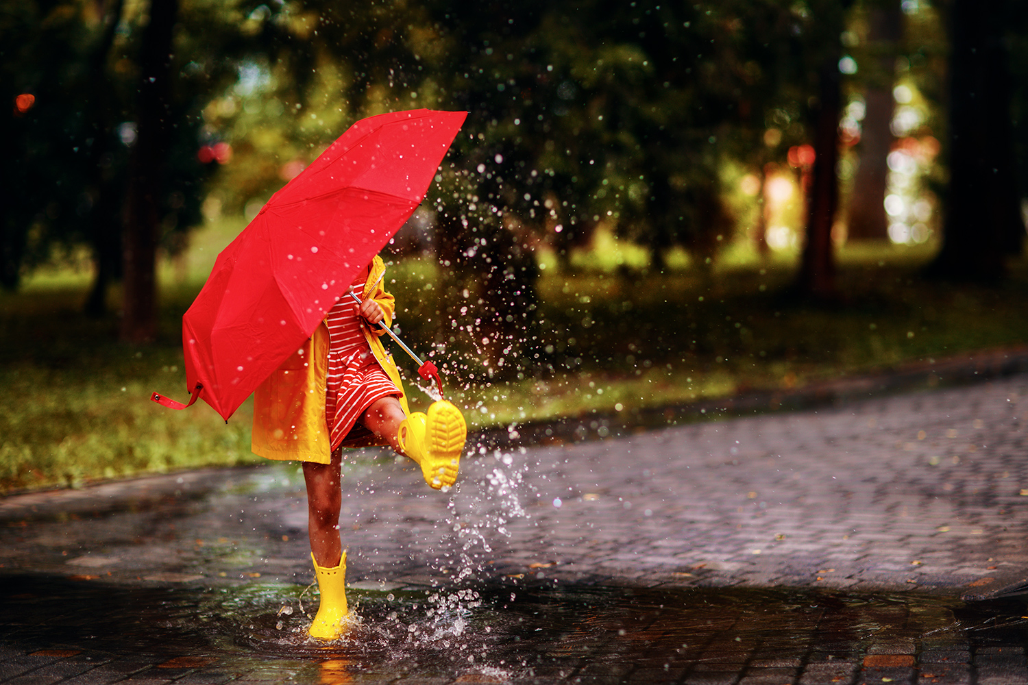 A child wearing boots and holding an umbrella splashes in a puddle.