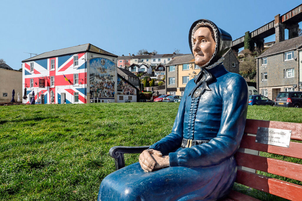 A statue of a woman in a blue dress and bonnet is seated on a bench and homes and other buildings are in the background.