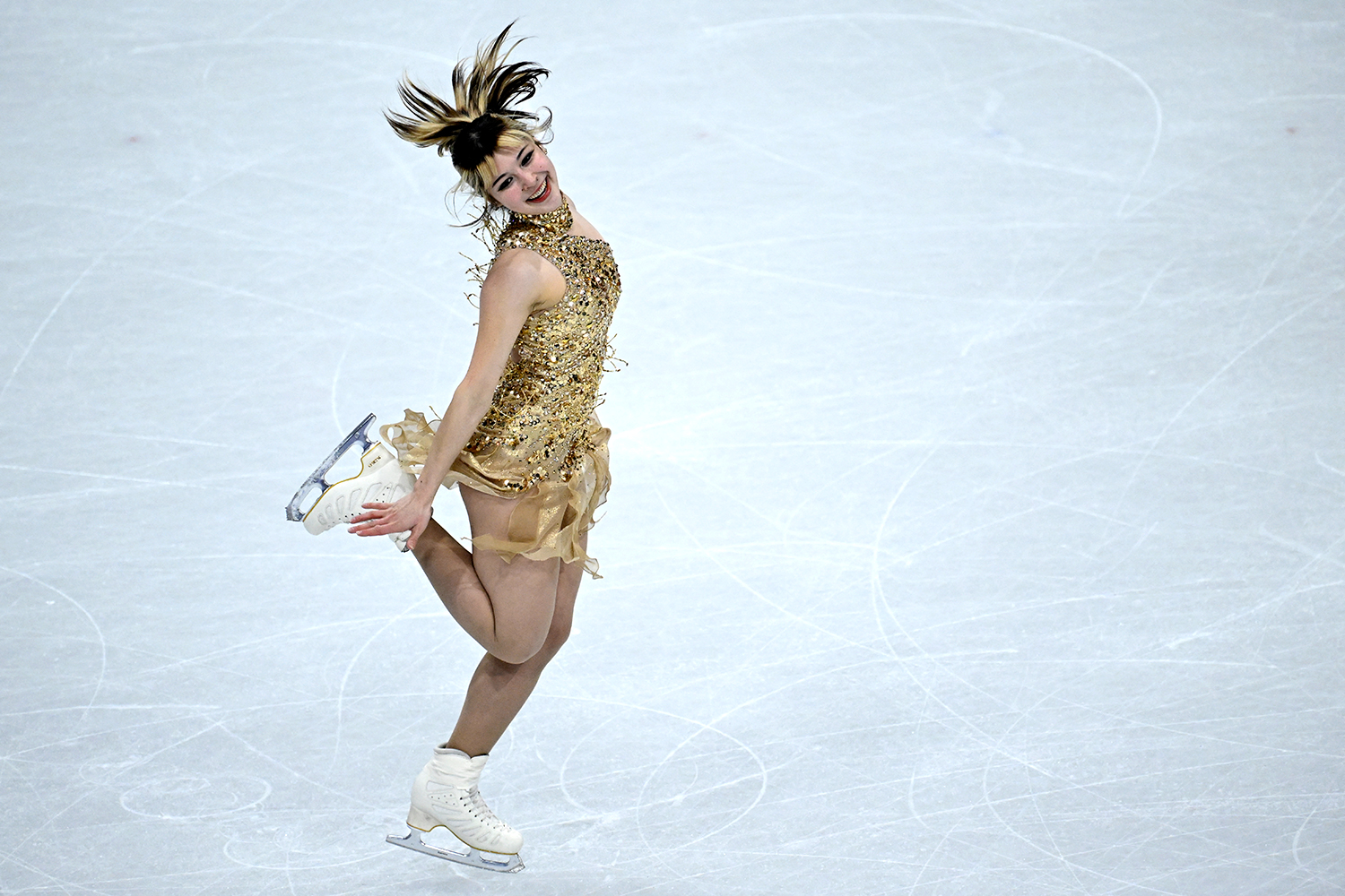 Alysa Liu stands on the ice and smiles while holding up leg in the air behind her.