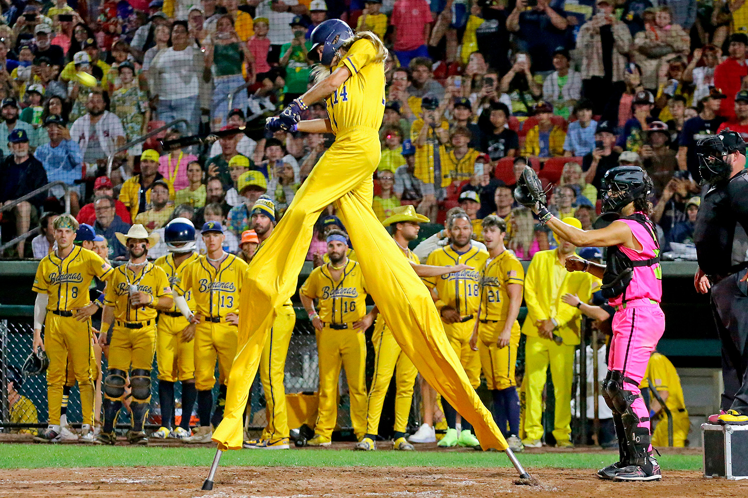 A baseball player on stilts in a bright yellow uniform is at bat in front of teammates and fans.