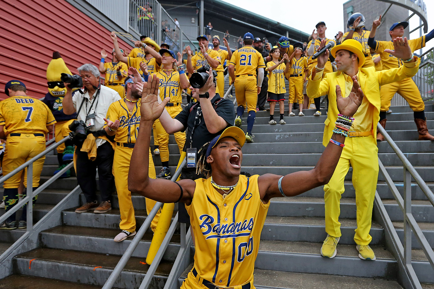 Savannah Banana players, along with Jesse Cole in a yellow suit, dance and clap their hands on stadium stairs.