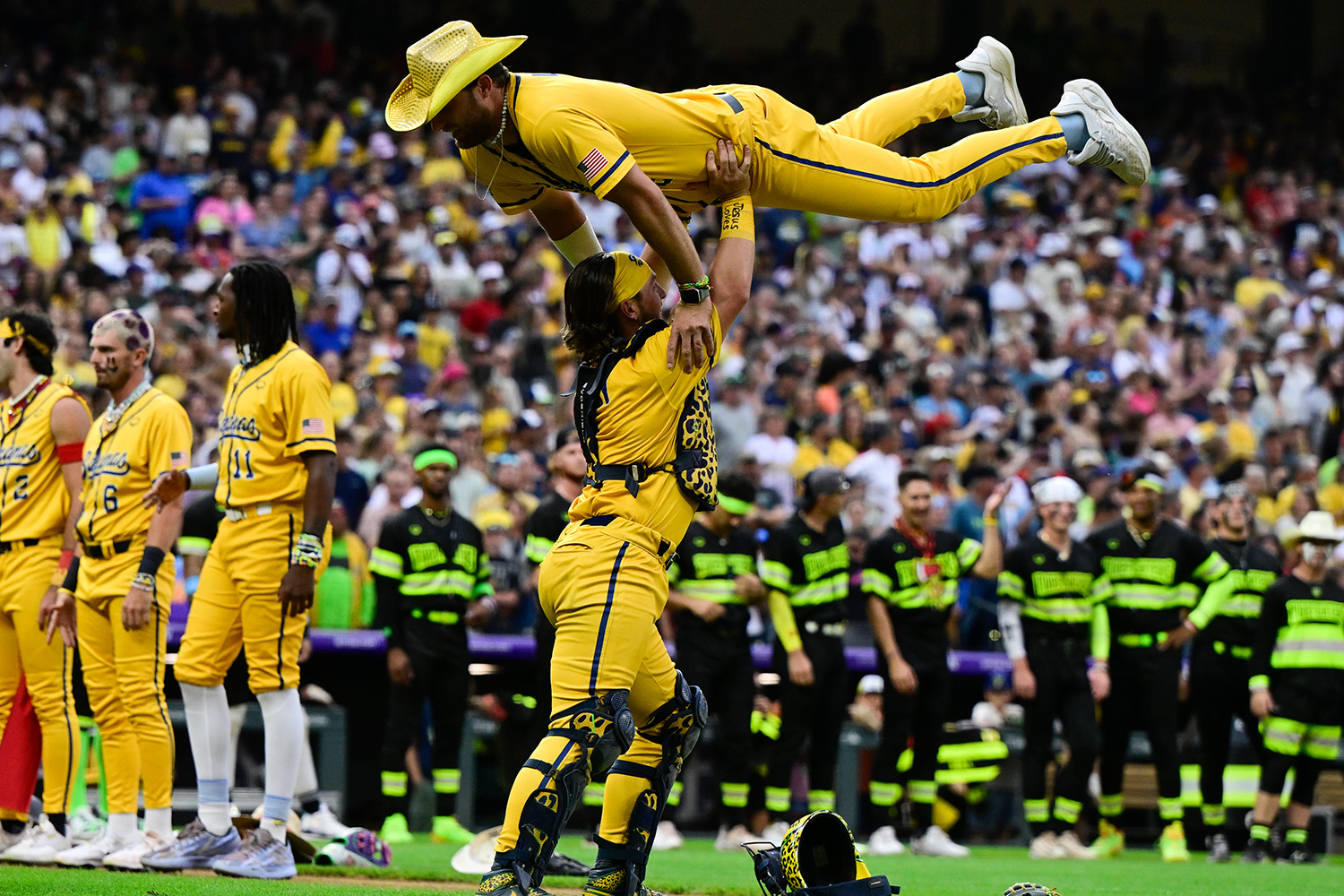 A member of the Bananas baseball team holds a teammate above his head as teammates and others watch.