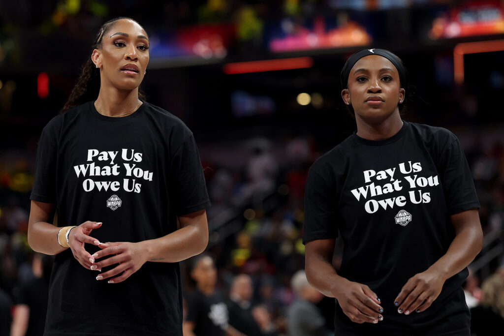 A’ja Wilson and Jackie Young stand on a basketball court wearing shirts that say “Pay us what you owe us.”