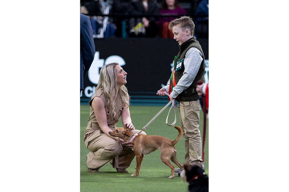 Owner Oliver Irwin stands with his small brown dog called Betty and talks to Dame Laura Kenny who is crouched and petting Betty.