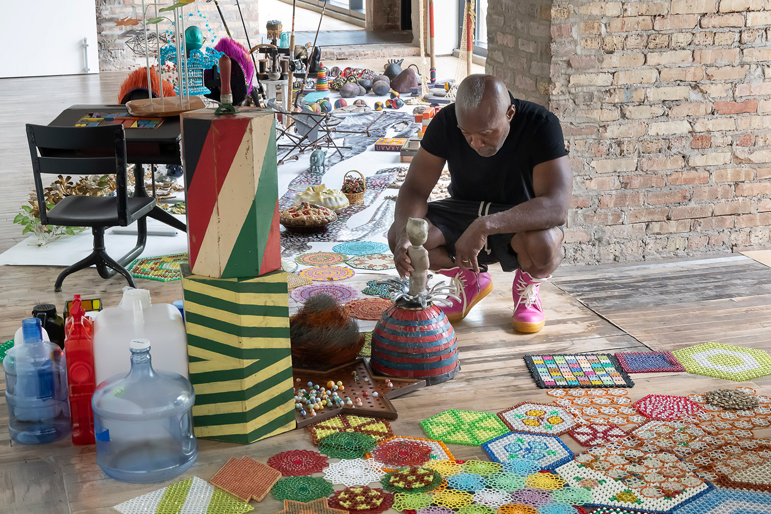Nick Cave crouches in his studio examining some of his art and surrounded by his other art as well as found objects.
