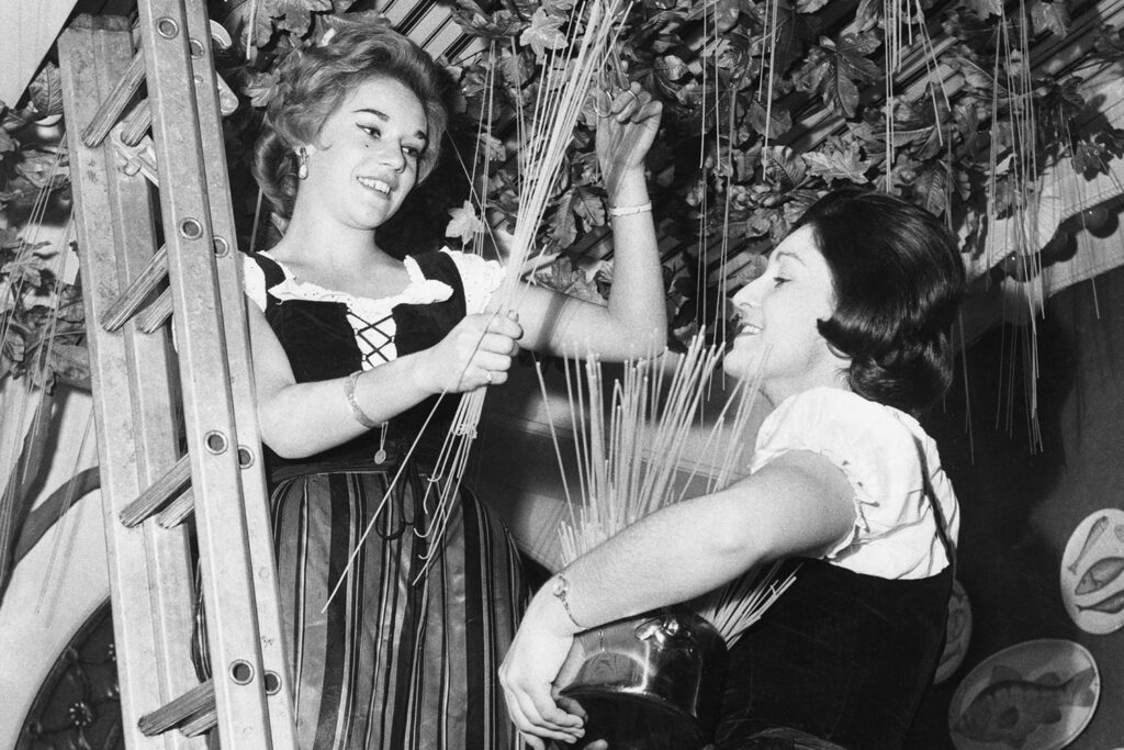 Two women appear to be cutting pieces of spaghetti that has been “growing” on a vine.