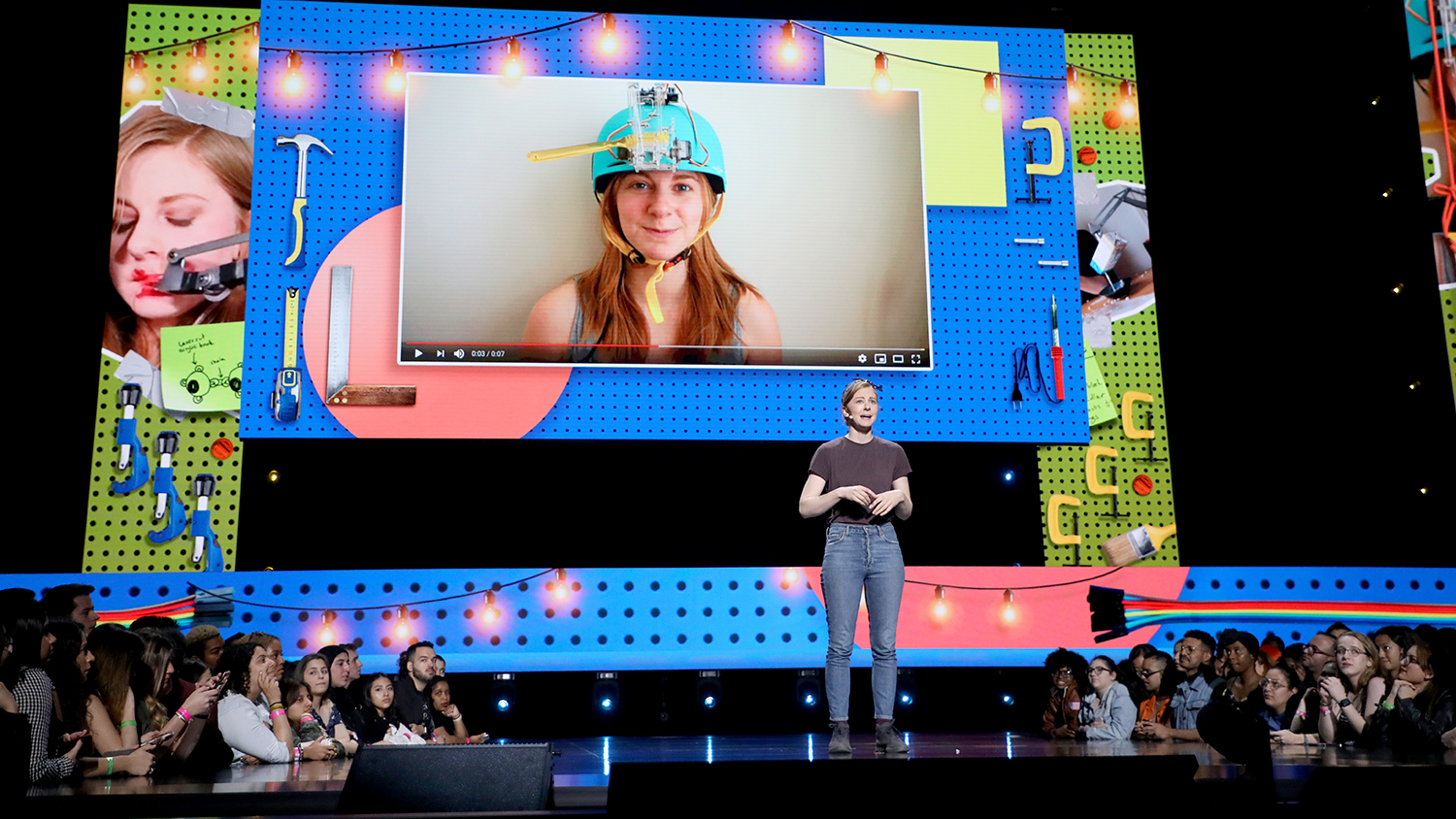 Simone Giertz stands on a stage in front of a screen showing a demonstration of her Toothbrush Machine.