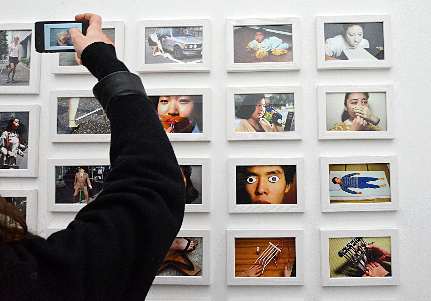 A hand holds up a phone to take a photo of a wall of photos of chindogu that are in use.