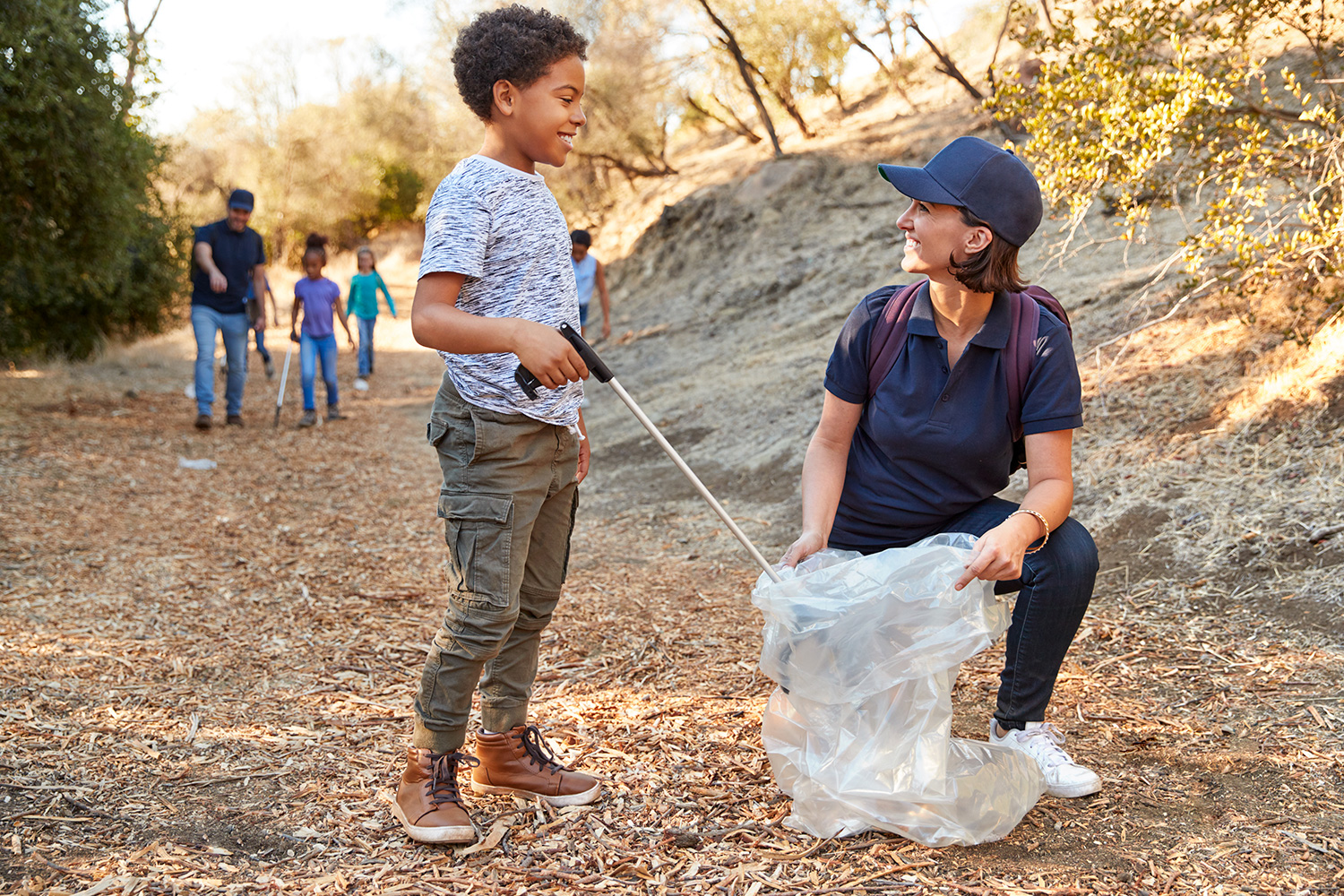 A child smiles as he puts trash into a plastic bag held by an adult, while other kids and adults look for trash behind them.
