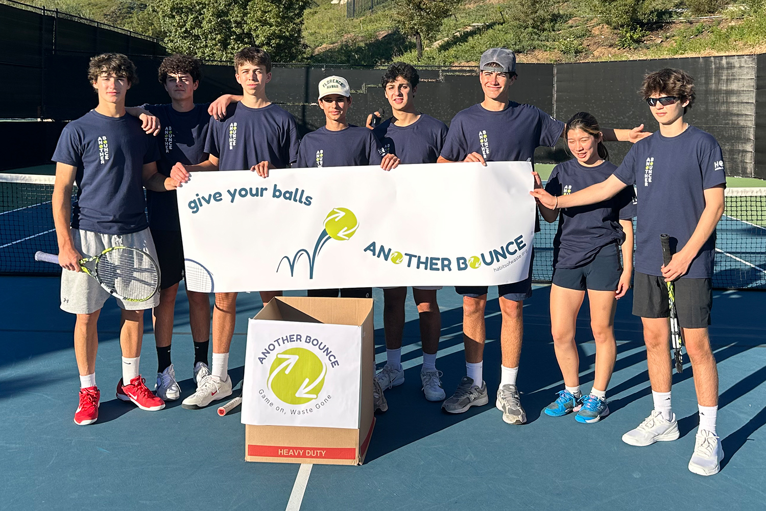 Eight teens pose on a tennis court with a sign that says Another Bounce.