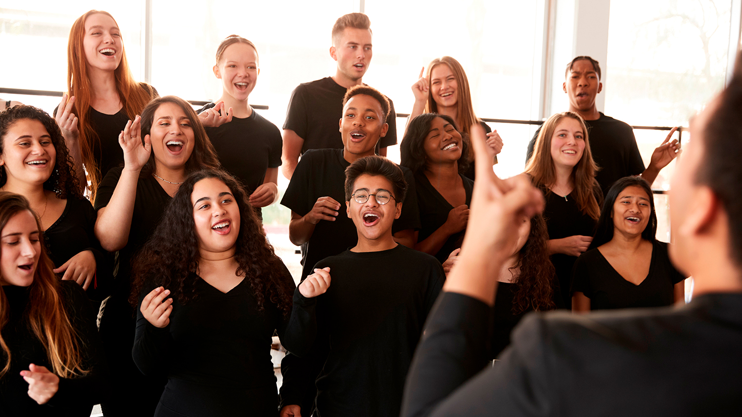 Teens or young adults smile and gesture as they sing in a choir with the help of a director.