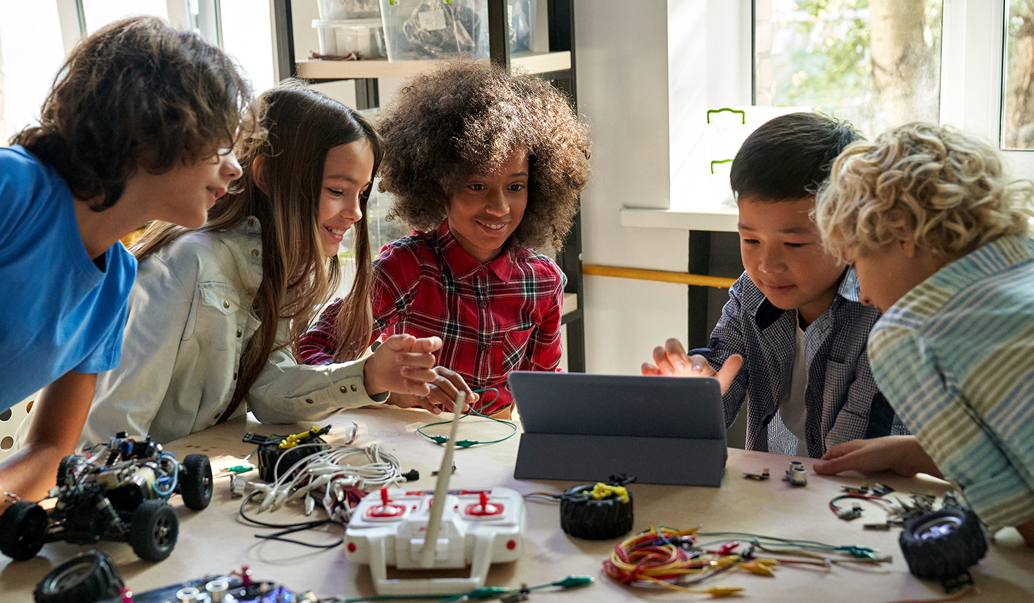 Children sit around a table and work on a computer and with robots equipment.