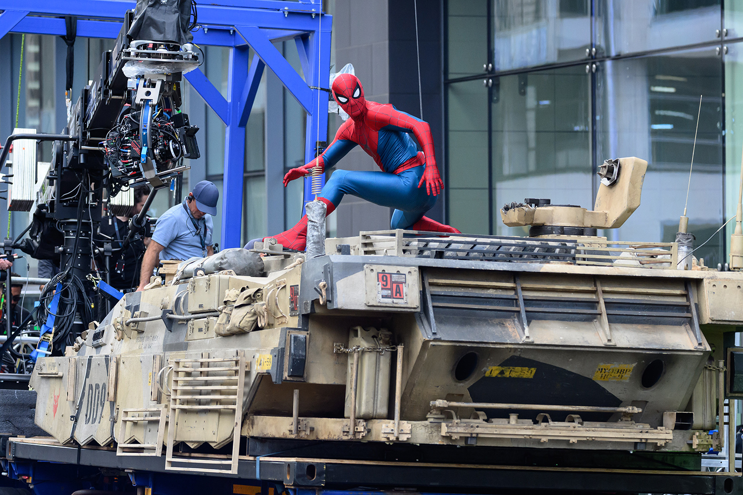 Tom Holland wears a Spiderman costume as he stands on a movie set with equipment and crew members.