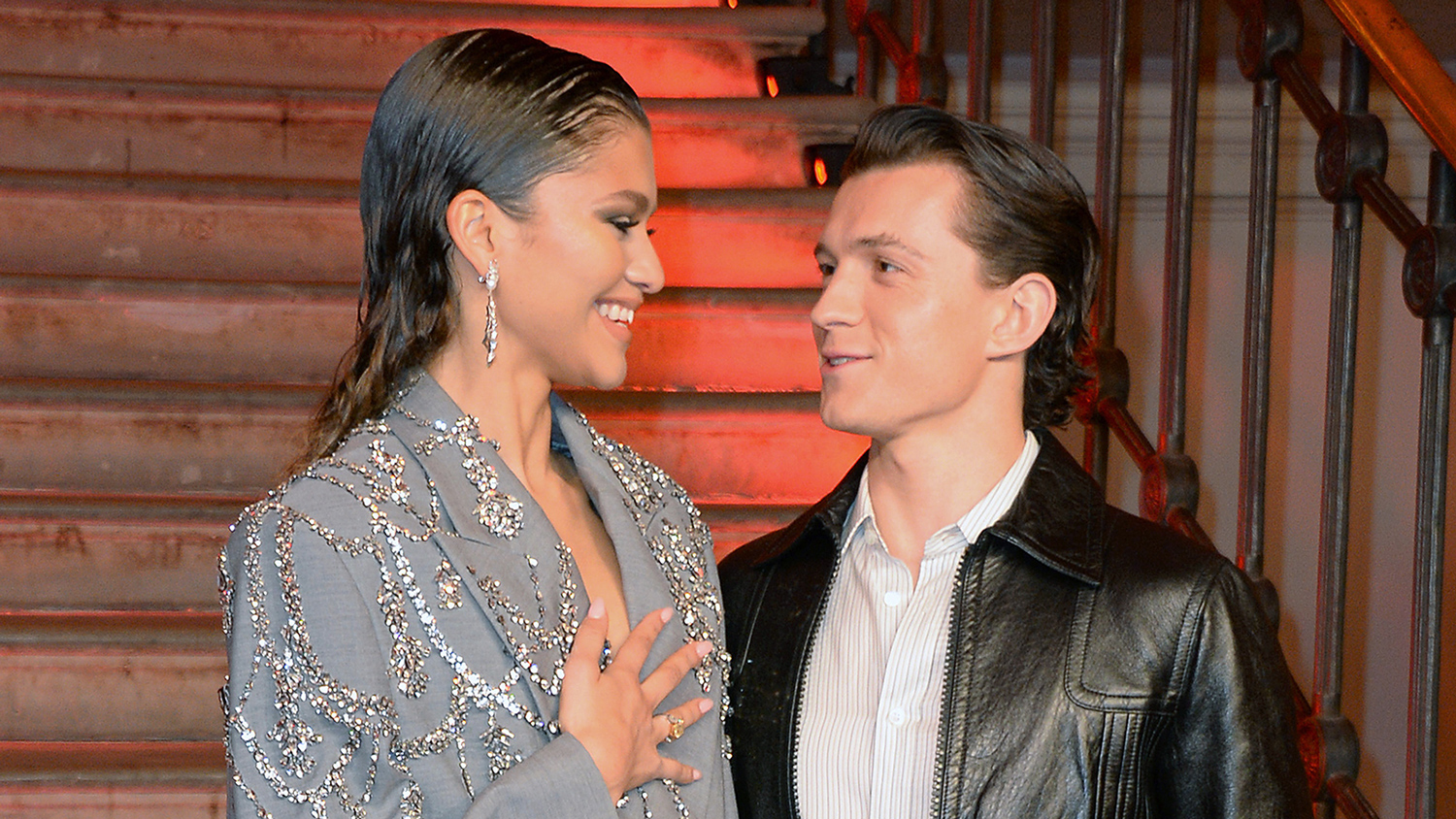 Zendaya and Tom Holland look at each other and smile as they pose in front of a staircase.