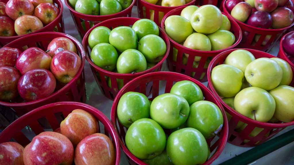 Four varieties of apples are in round red baskets at a market.