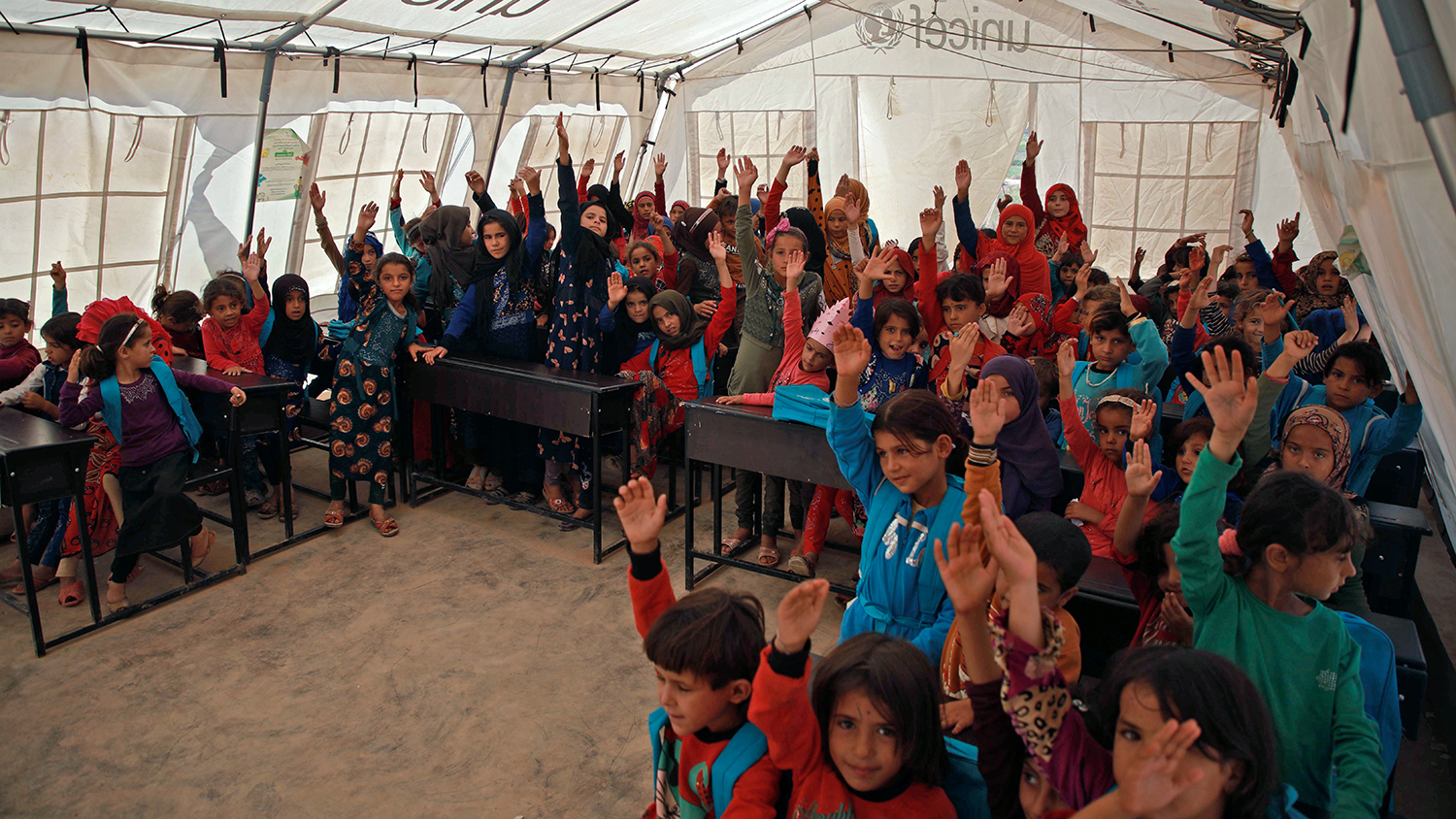 Young children sit at desks inside a UNICEF tent. Each child has a hand raised.