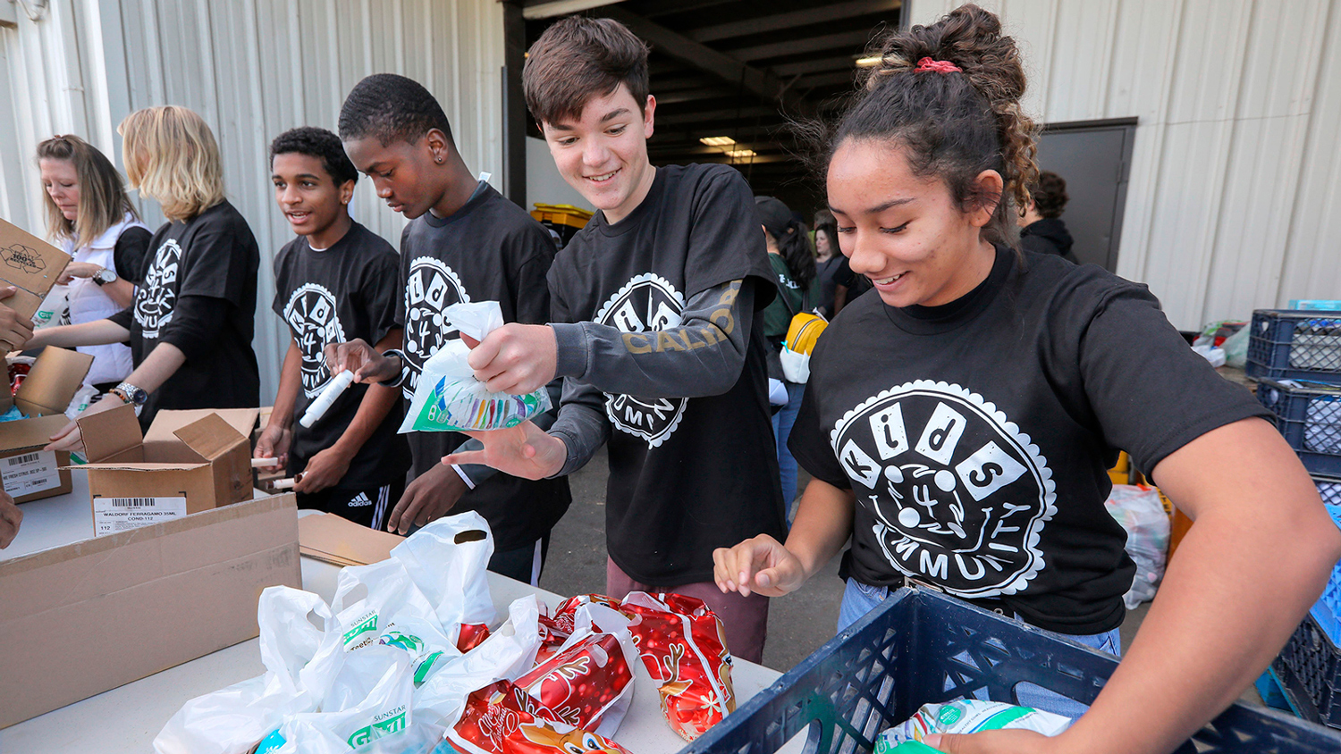 Five young people wearing Kids4Community shirts stand at an outdoor table putting items into plastic bags.