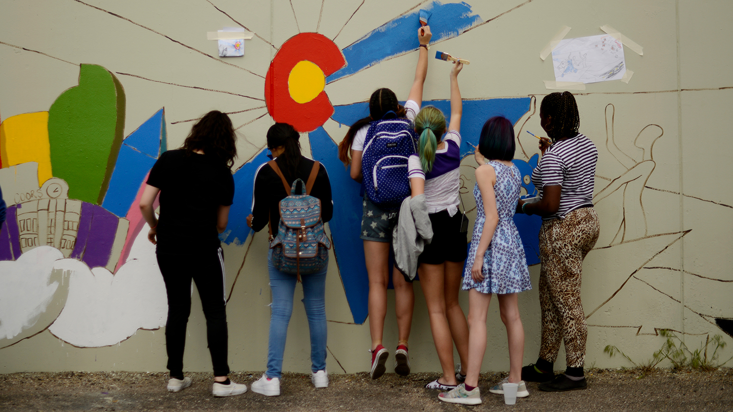 Six teens have their backs to the camera as they paint a wall mural.