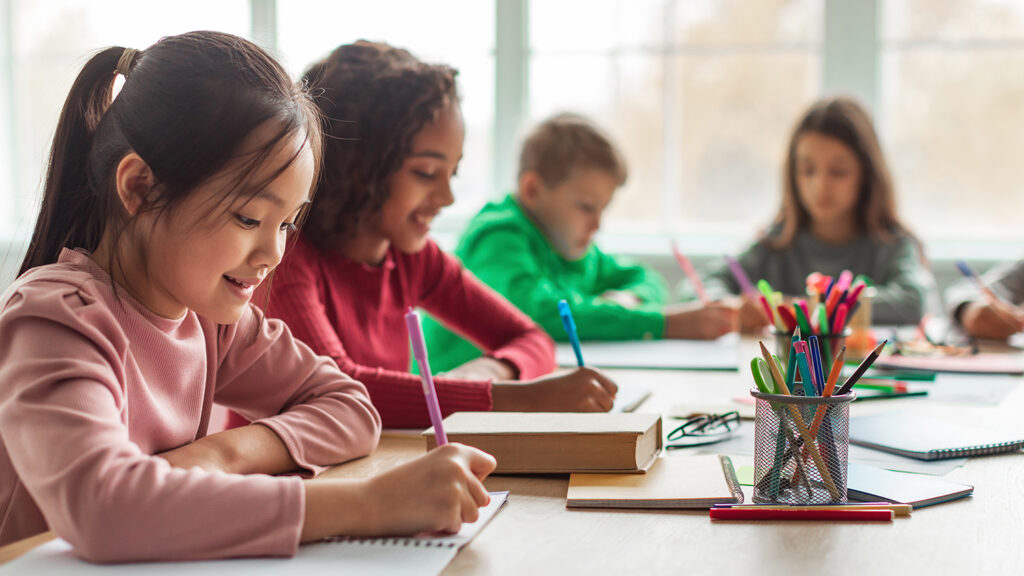 Five students sit around a table using pens to write on paper.