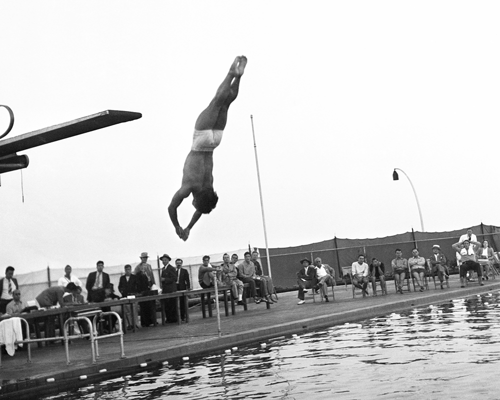 Sammy Lee is mid-dive over an outdoor pool as several people watch from the side.