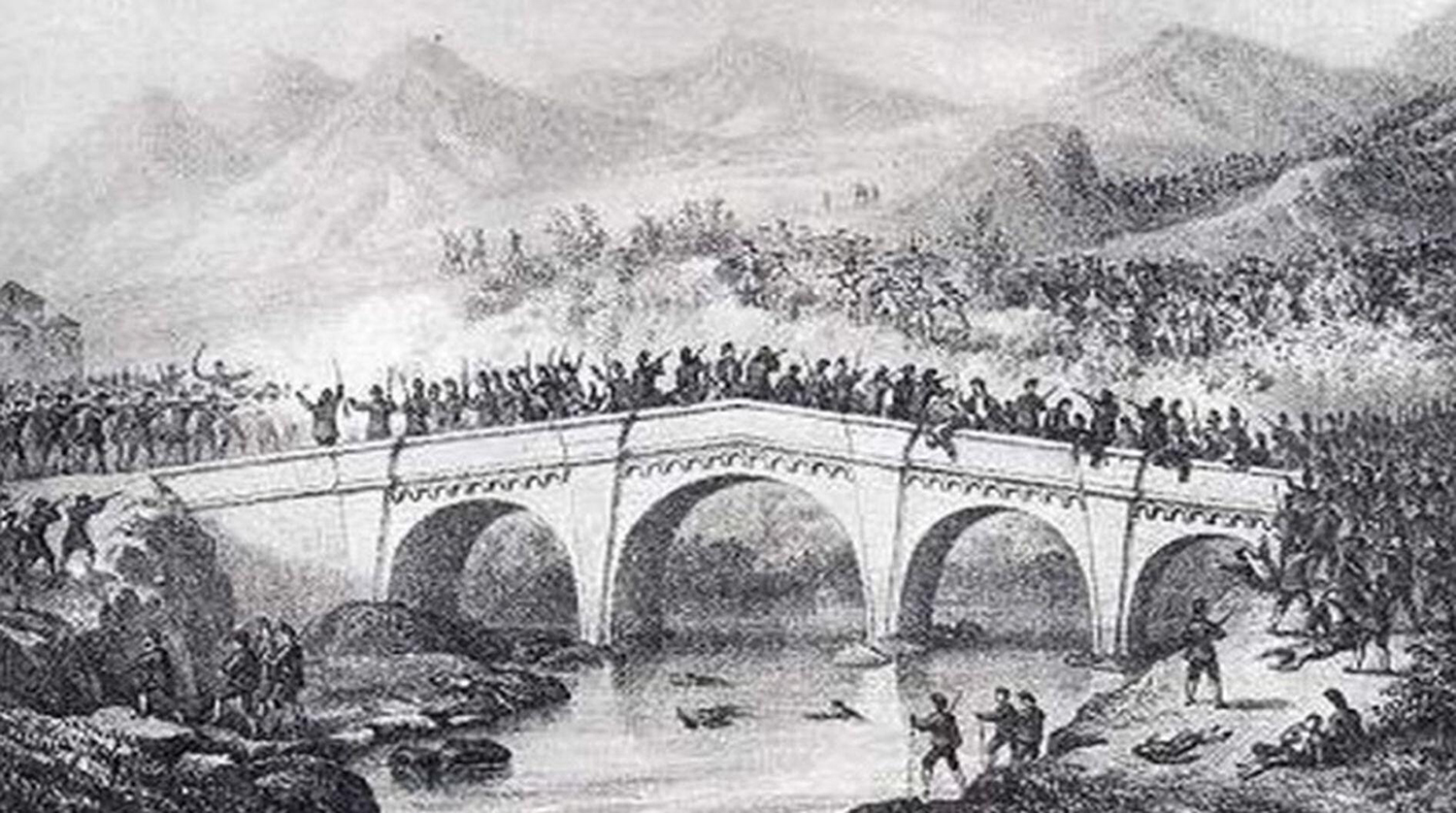 Soldiers do battle on and around a bridge with mountains in the background.