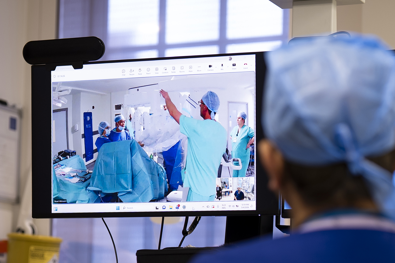The back of a surgeon’s head is seen looking at a screen on which doctors are prepping for surgery in another location.
