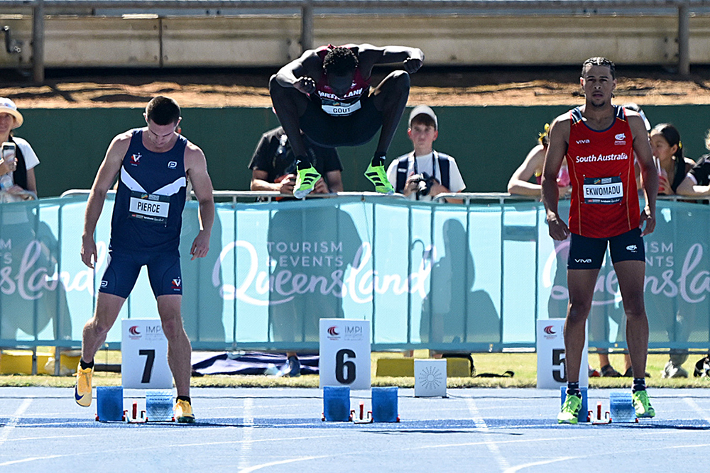 Gout Gout jumps high into the air as runners on each side of him stand at their starting blocks.