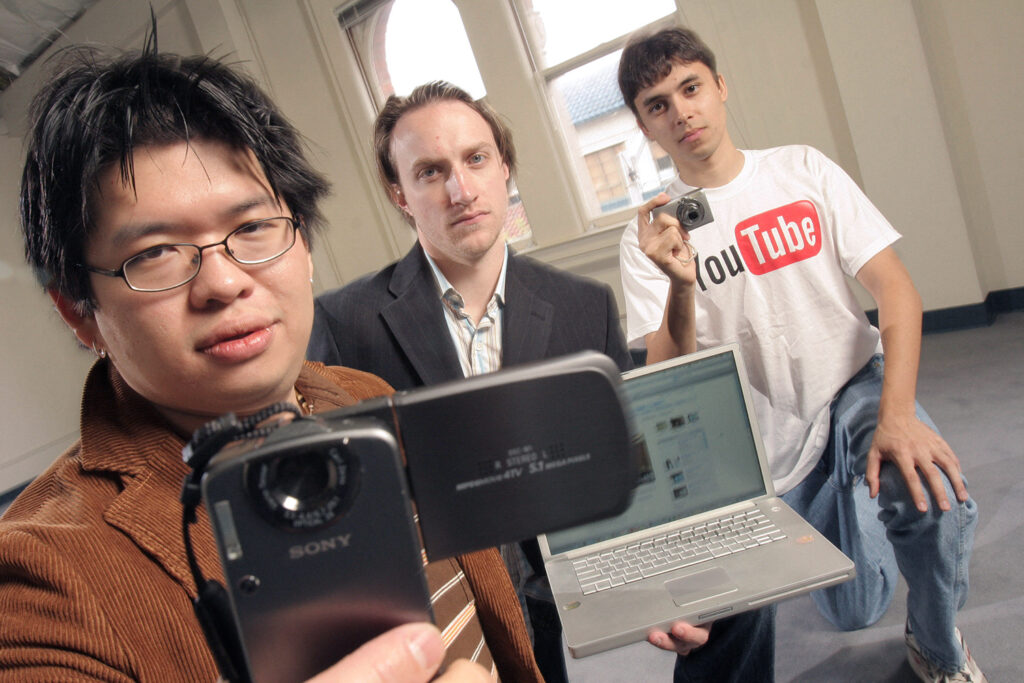 Steve Chen, Chad Hurley, and Jawed Karim pose in a large room while holding a video recorder, a laptop, and a camera.