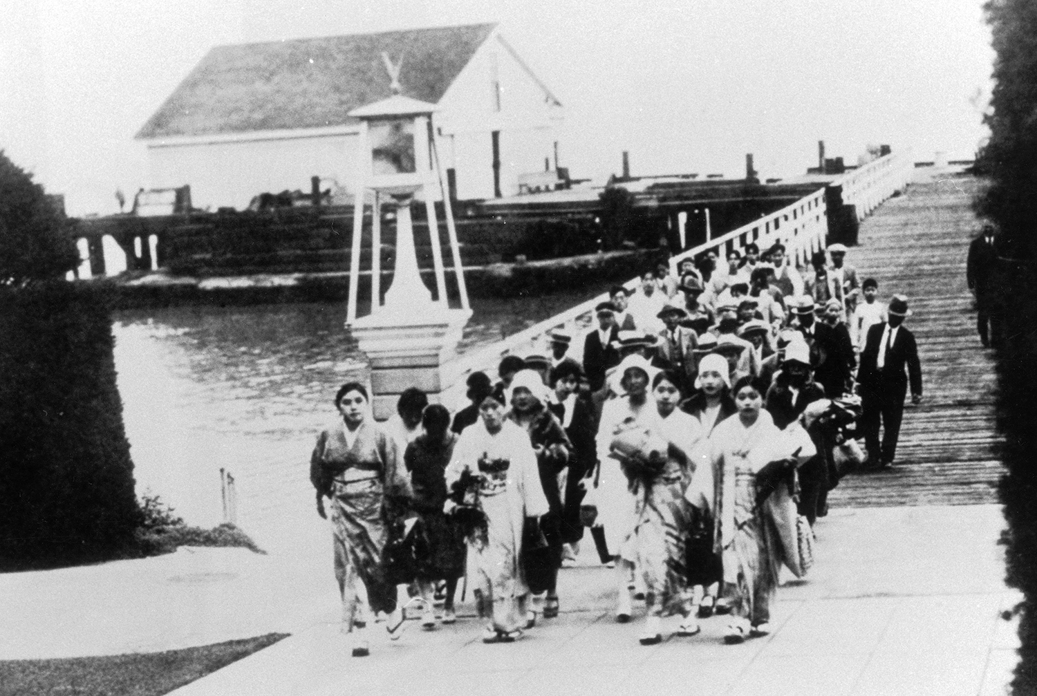 Asian immigrants in early 20th century clothing walk across a bridge over a body of water.